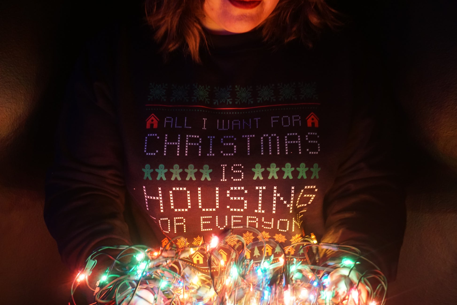 a person holding a string of lights while wearing a navy crewneck sweater that has 'all I want for christmas is housing for everyone' in a cross-stitch style design on the front